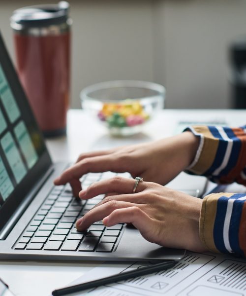 Unknown woman working on laptop, designing mobile application interface, hands typing on keyboard, digital wireframes visible on screen, office desk in background