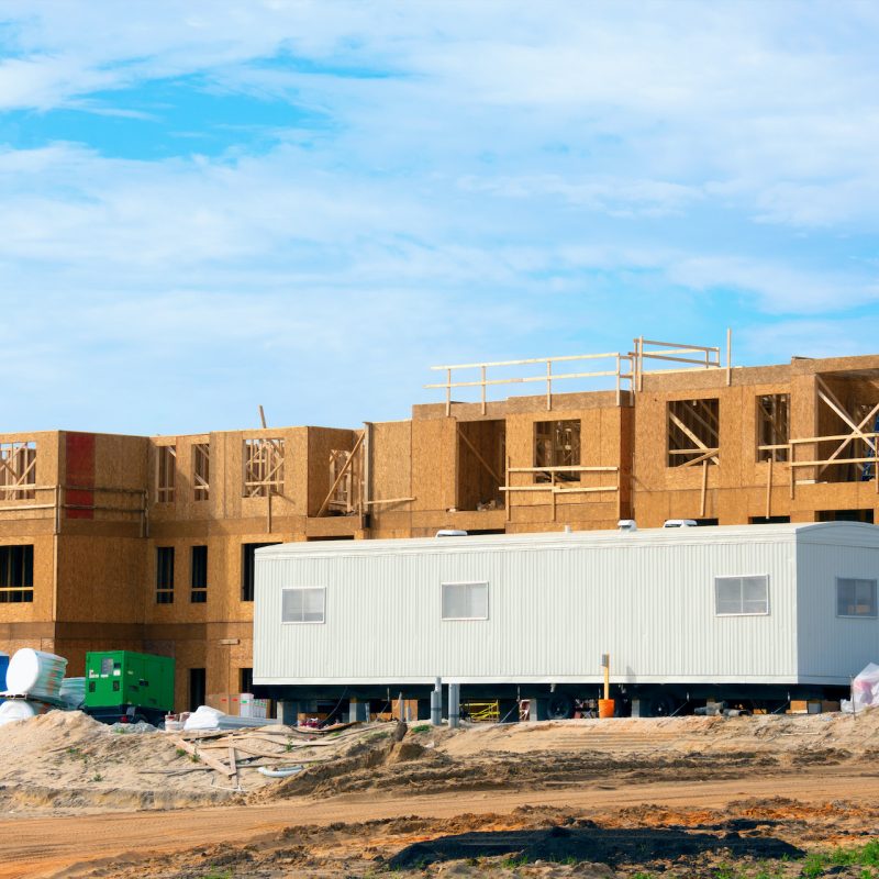 Large construction site half constructed wooden building with mobile job location office trailer in front area for contractors, work managers and supervisors for paperwork and worker interviews.