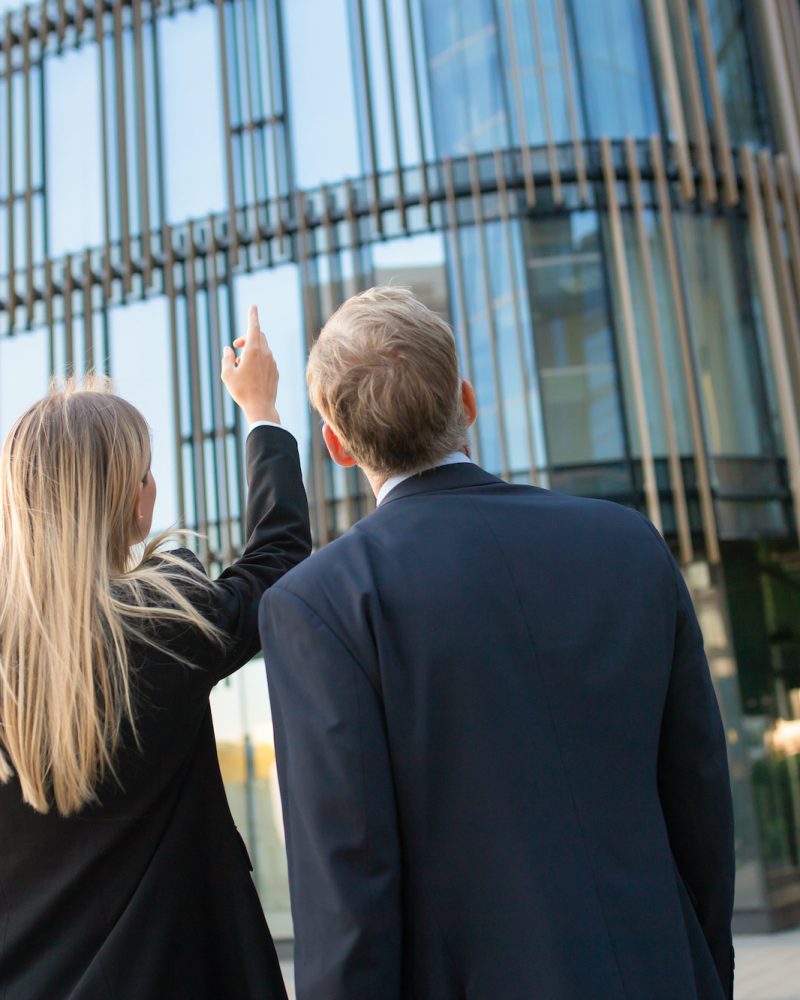 Business professionals in suits looking at office building facade, pointing up, discussing real property. Back view, low angle. Commercial real estate concept