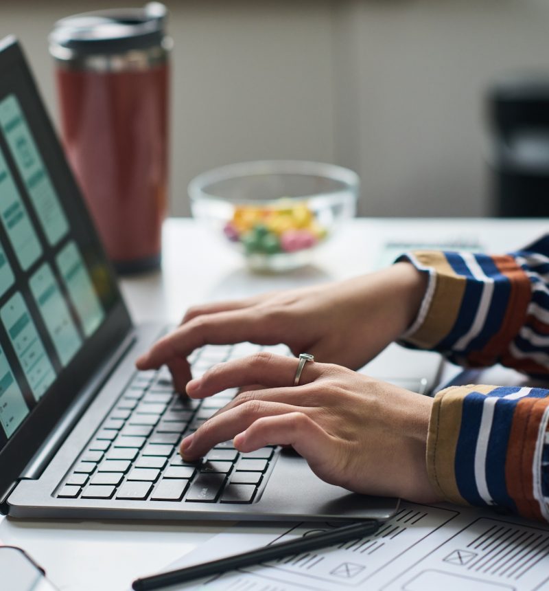 Unknown woman working on laptop, designing mobile application interface, hands typing on keyboard, digital wireframes visible on screen, office desk in background